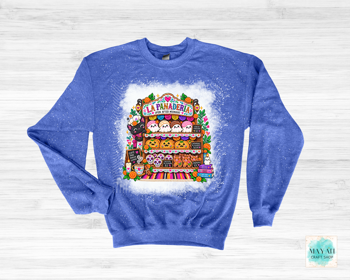 A royal blue crewneck sweatshirt with a white paint-splatter effect, featuring a colorful graphic of a Halloween-themed 'La Panadería' bakery stall with cute cartoon ghosts. The sweatshirt is laid flat on a light gray wood-plank background.
