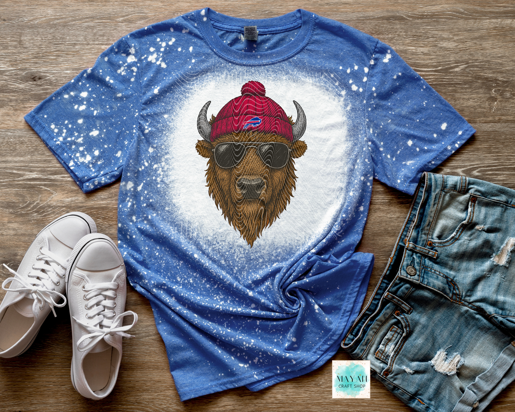 Blue t-shirt with a graphic of a bison wearing sunglasses and a beanie, paired with white sneakers and denim shorts on a wooden surface.