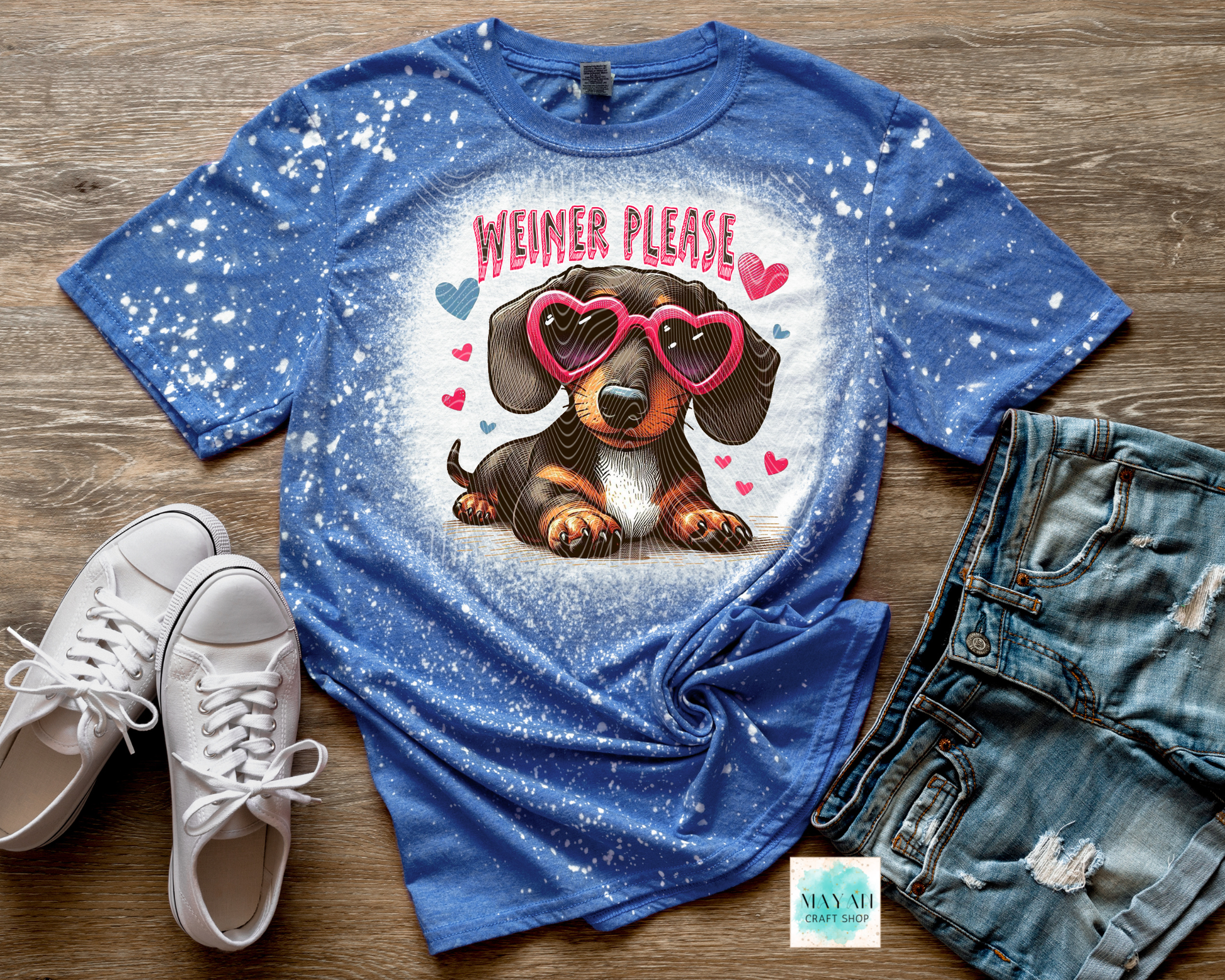 Blue t-shirt with dachshund graphic and 'Weiner Please' text on a wooden surface with shoes and shorts.