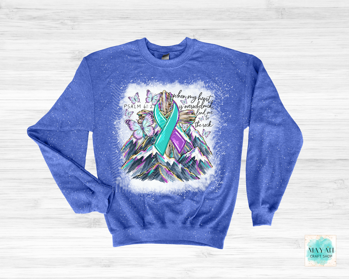 A heathered blue crewneck sweatshirt featuring a colorful graphic of a mountain with butterflies and a teal and purple awareness ribbon, laid flat on a white wood background.