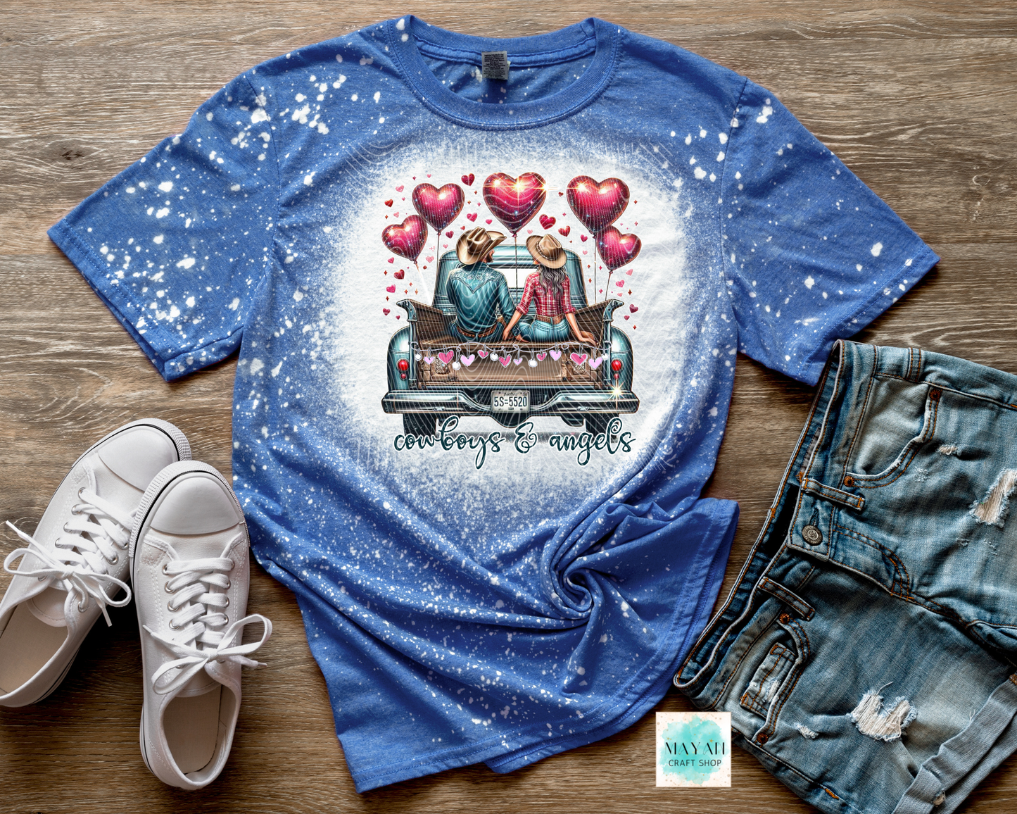 Blue t-shirt with a graphic design of two people in a car with hearts, paired with white sneakers and denim shorts on a wooden surface.