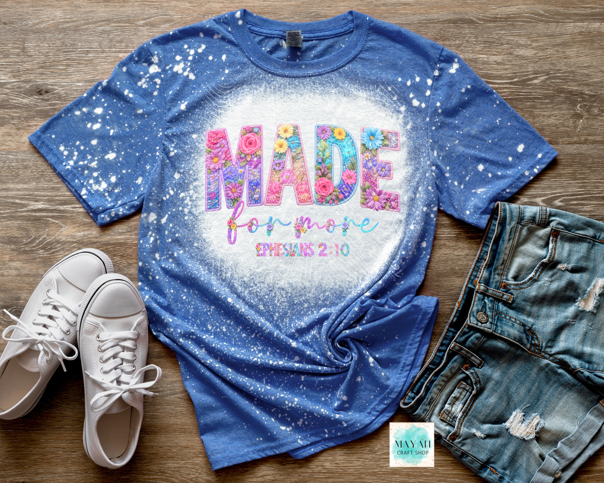 Blue t-shirt with colorful text and floral design, paired with white sneakers and denim shorts on a wooden surface.