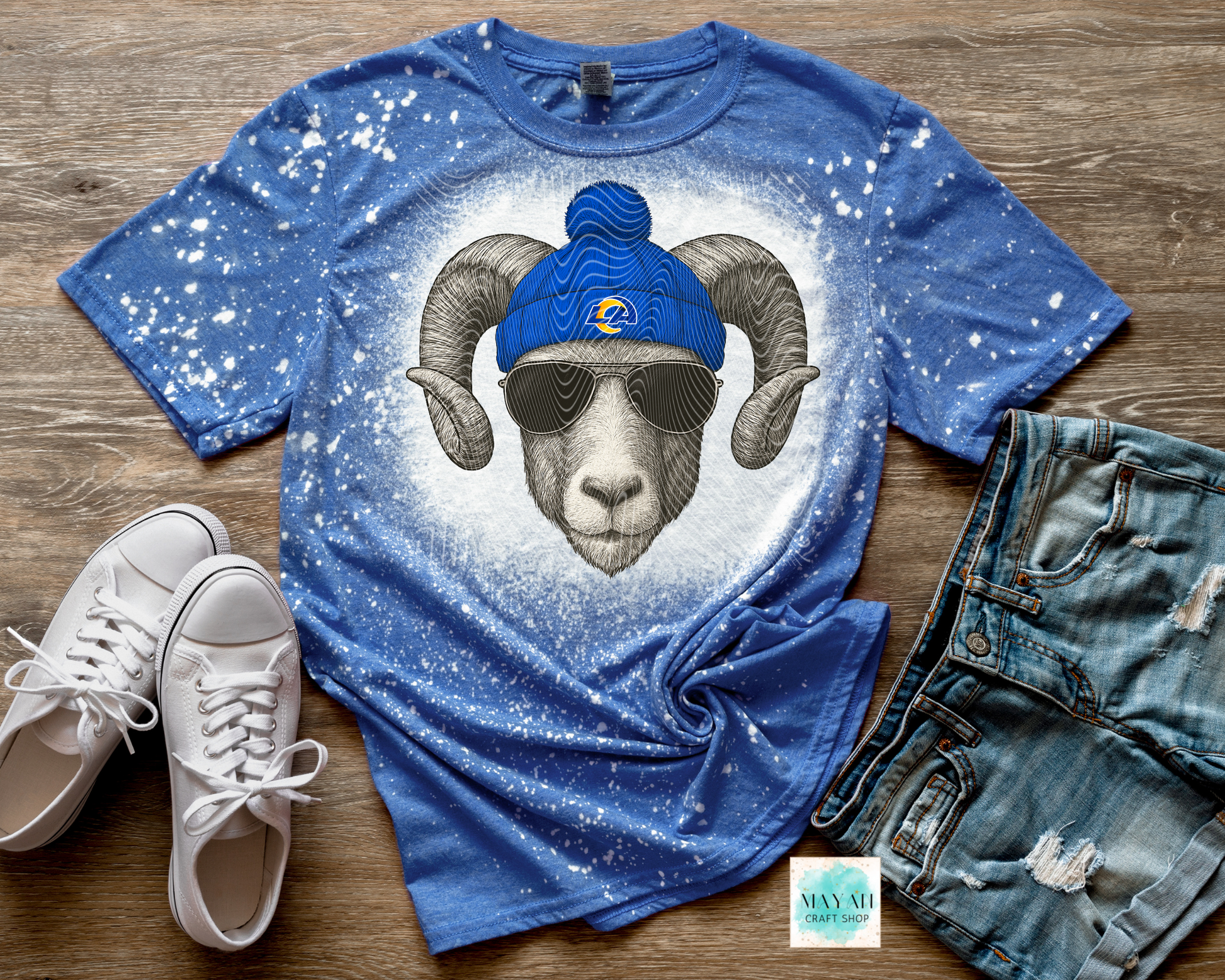 Blue t-shirt with a graphic of a ram wearing sunglasses and a blue cap, paired with white sneakers and denim shorts on a wooden surface.