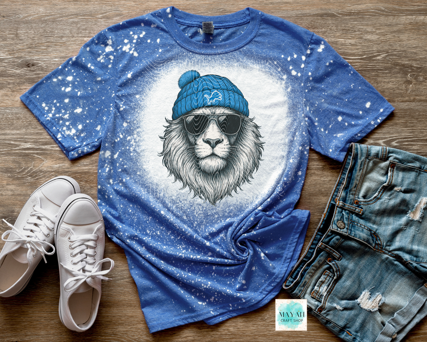 Blue t-shirt with a lion graphic wearing a blue beanie and sunglasses, paired with white sneakers and denim shorts on a wooden surface.