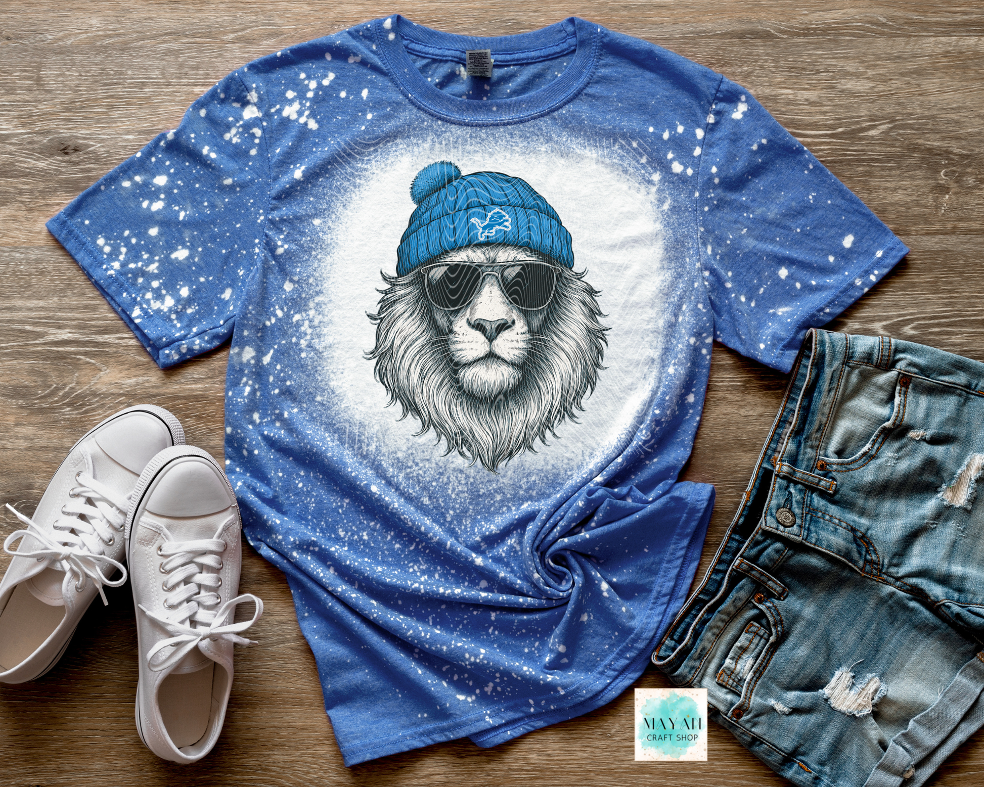 Blue t-shirt with a lion graphic wearing a blue beanie and sunglasses, paired with white sneakers and denim shorts on a wooden surface.