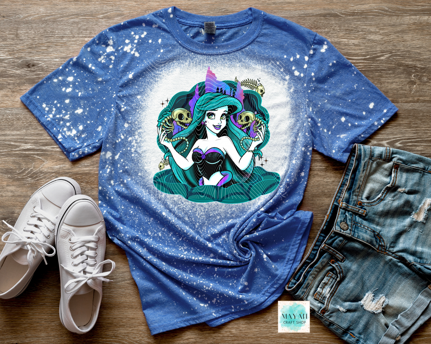 Blue t-shirt with a colorful graphic design of a mermaid, paired with white sneakers and denim shorts on a wooden surface.