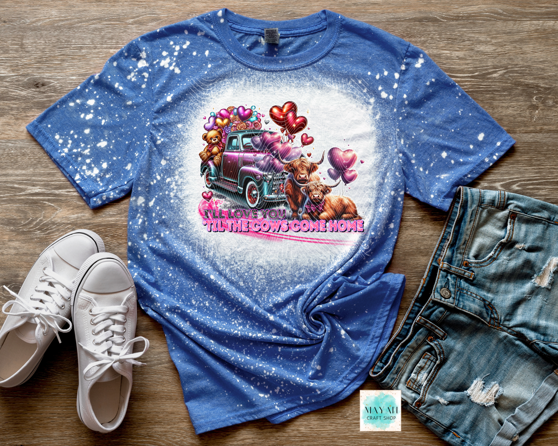 Blue t-shirt with a graphic design of a pink truck and cow, paired with white sneakers and denim shorts on a wooden surface.