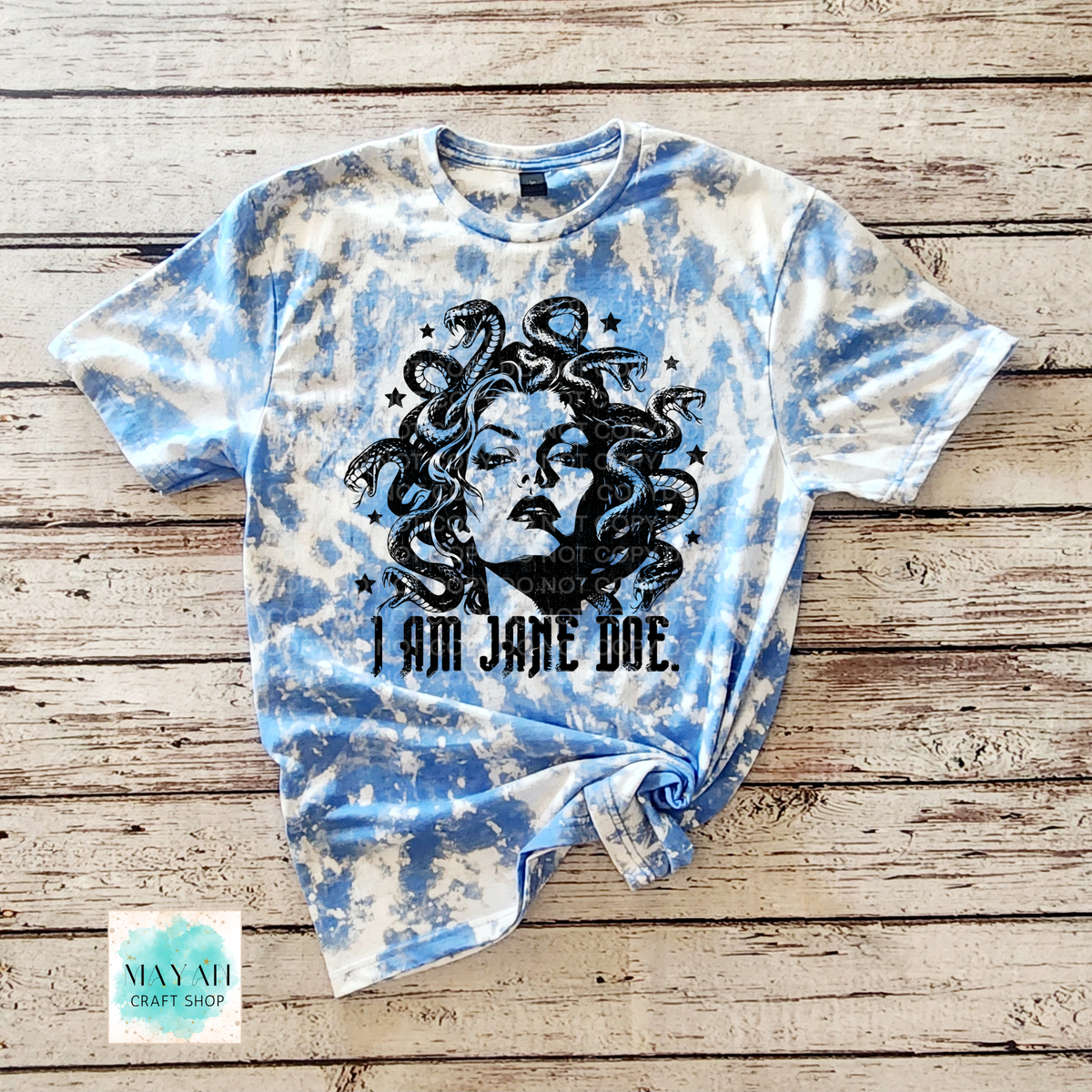 A heather royal blue bleached graphic t-shirt featuring a black illustration of Medusa's face with flowing hair and the text 'I AM JANE DOE' printed below it. The t-shirt is laid flat on a rustic wooden plank background with a 'May Mu Craft Shop' logo in the bottom-left corner.