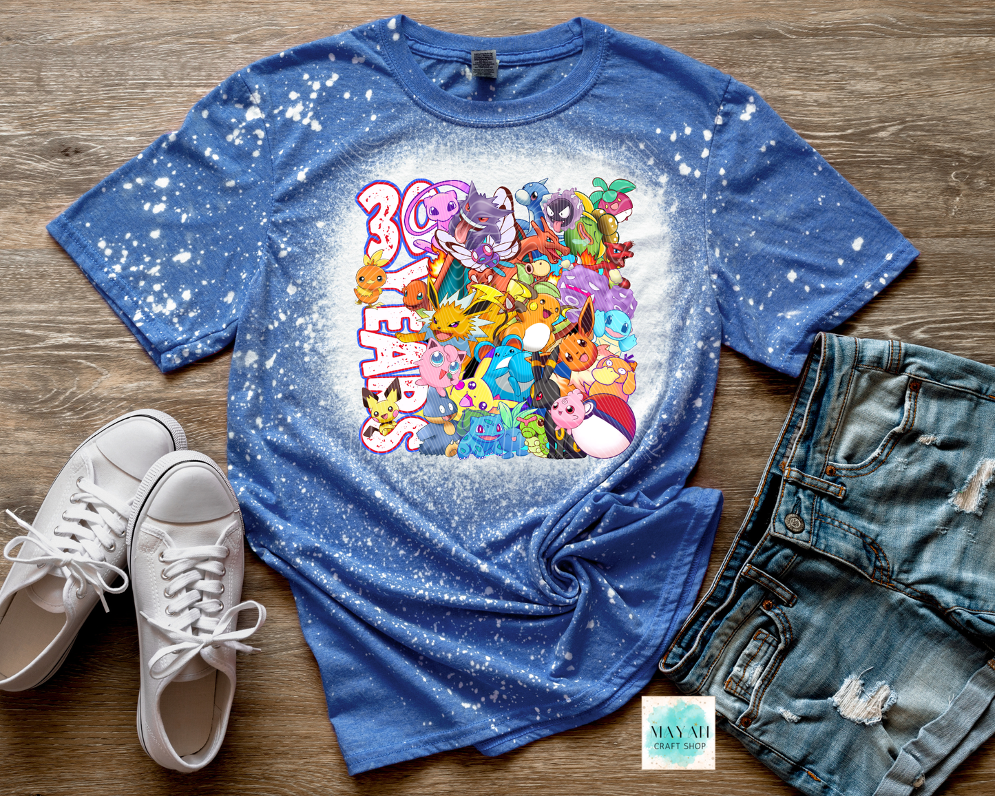 Blue t-shirt with colorful graphic design on a wooden surface with sneakers and jeans.