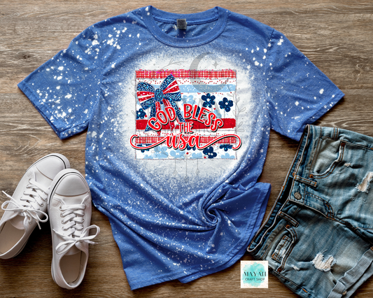 Blue t-shirt with 'God Bless the USA' graphic on a wooden surface with white sneakers and denim shorts.