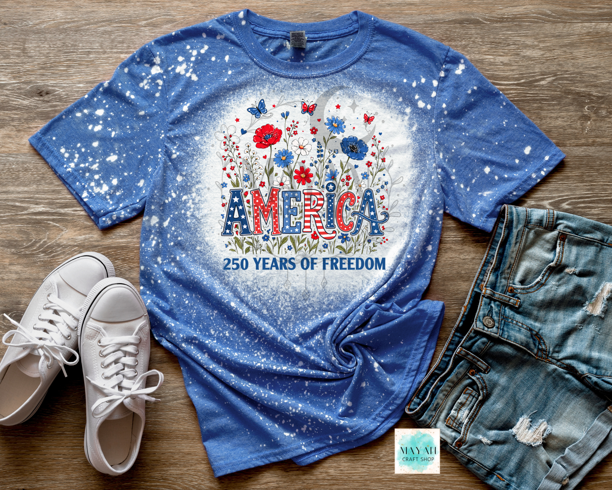 Blue t-shirt with 'America 250 Years of Freedom' design on a wooden surface with white sneakers and denim shorts.