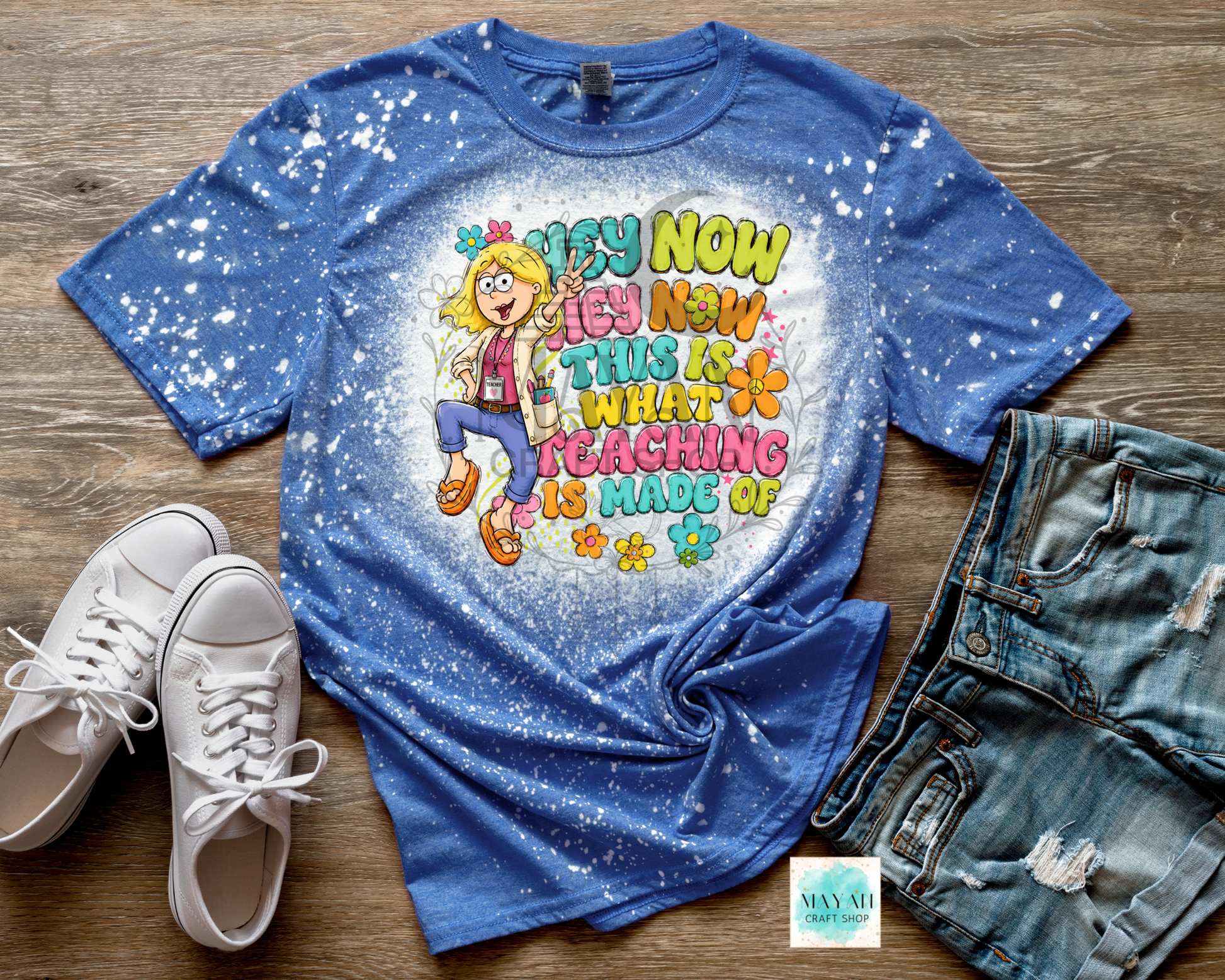 A blue splatter-dyed graphic t-shirt featuring a cartoon teacher and the text 'I'M NOW I'M NOW THIS IS WHY TEACHING IS MADE OF FUN' in colorful letters. The t-shirt is styled in a flat lay on a wooden floor with white sneakers on the left and distressed denim shorts on the right.