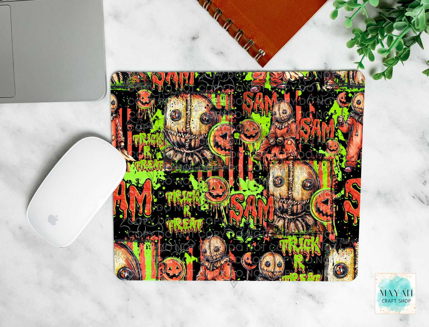 A mouse pad with a Halloween-themed graphic print featuring jack-o'-lanterns, dripping green slime, and spooky faces on a black background. The mouse pad is shown in a flat-lay setting on a white marble surface next to a white computer mouse, part of a silver laptop, an orange spiral notebook, and a green plant.