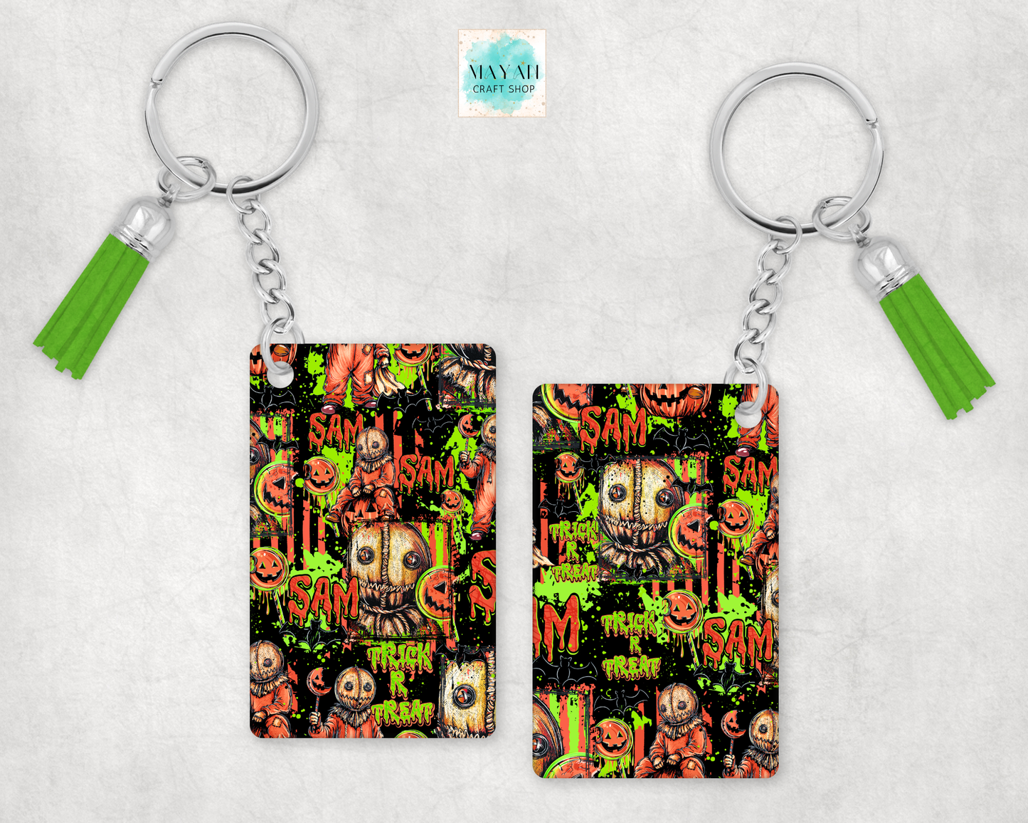 Two keychains with silver key rings and bright green tassels, featuring a Halloween-themed graphic print with pumpkins, skulls, and a word on a light gray textured background.