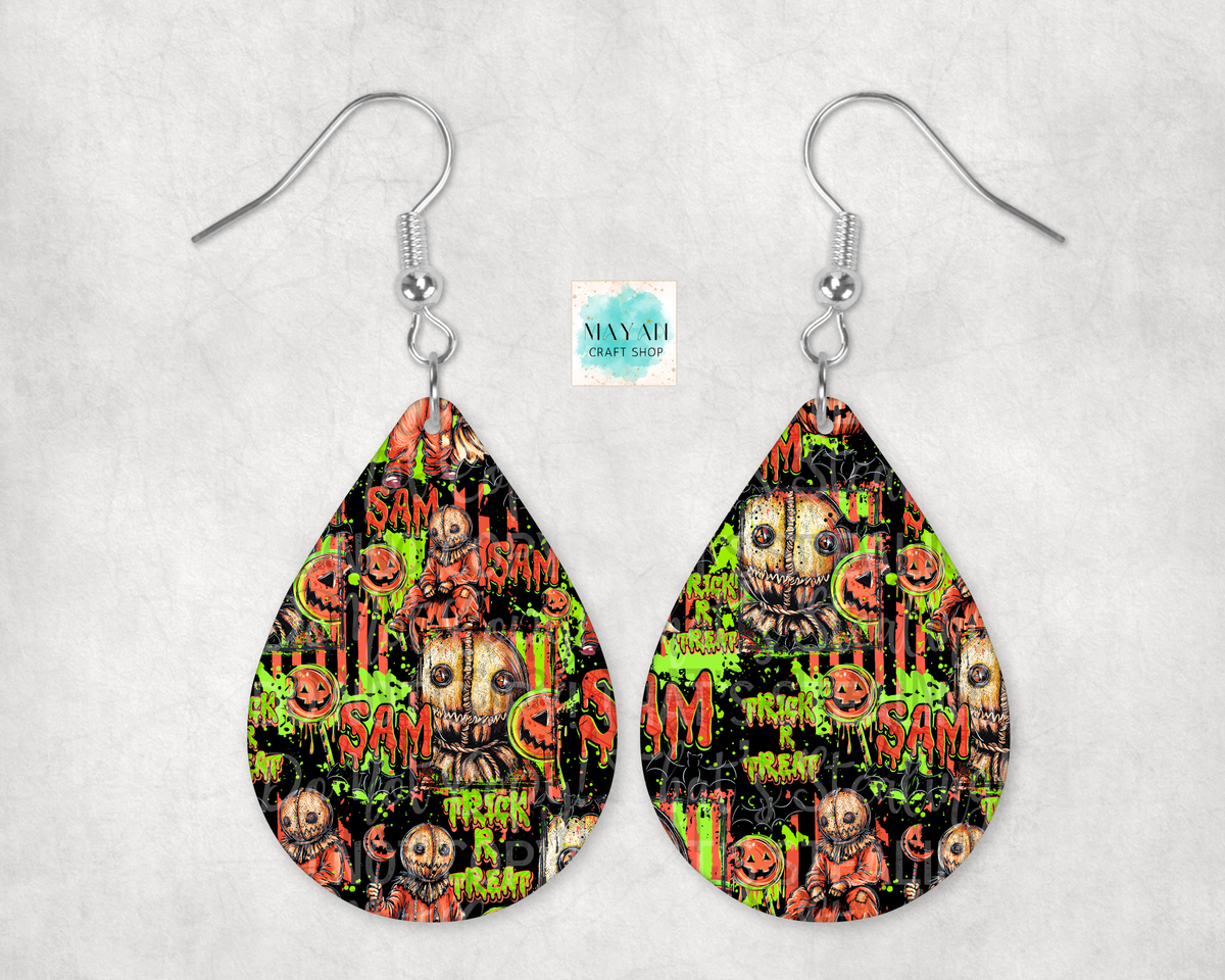 A pair of teardrop-shaped dangle earrings with a colorful Halloween-themed graphic print featuring skulls, pumpkins, suspended from silver fishhook wires against a light gray textured backdrop.