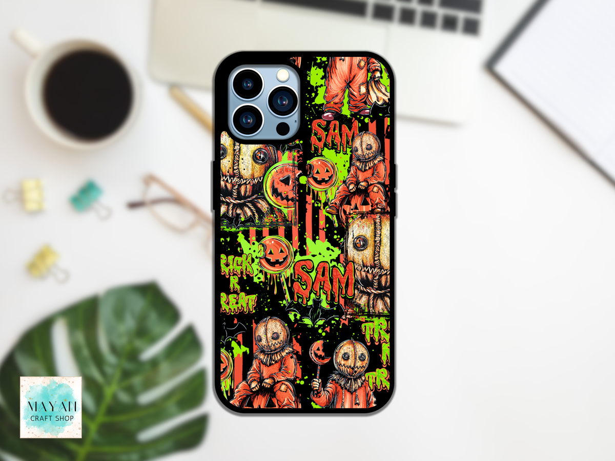 A phone case featuring a Halloween-themed collage print with jack-o'-lanterns, scarecrows, and words, shown on a white desk with a laptop, coffee cup, and leaf in the background.