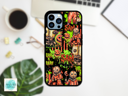 A phone case featuring a Halloween-themed collage print with jack-o'-lanterns, scarecrows, and words, shown on a white desk with a laptop, coffee cup, and leaf in the background.
