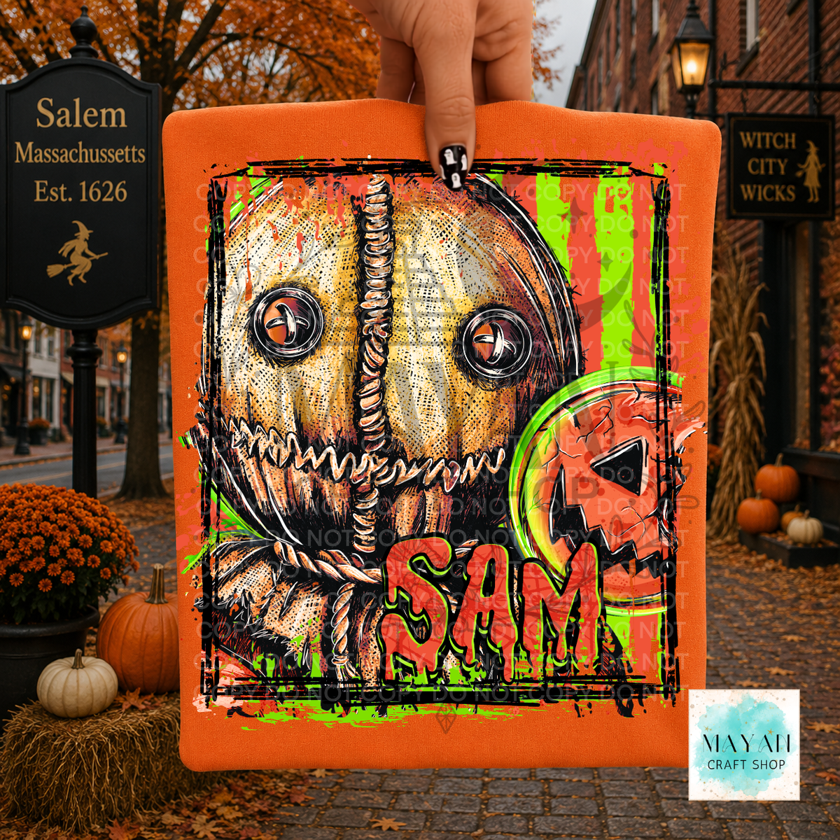 A hand holds a square orange canvas print featuring a horror-themed pumpkin design with a text and a witch hat motif. The background shows a real outdoor autumn street scene with Halloween decorations, pumpkins, and storefront signs.