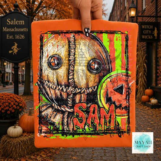 A hand holds a square orange canvas print featuring a horror-themed pumpkin design with a text and a witch hat motif. The background shows a real outdoor autumn street scene with Halloween decorations, pumpkins, and storefront signs.