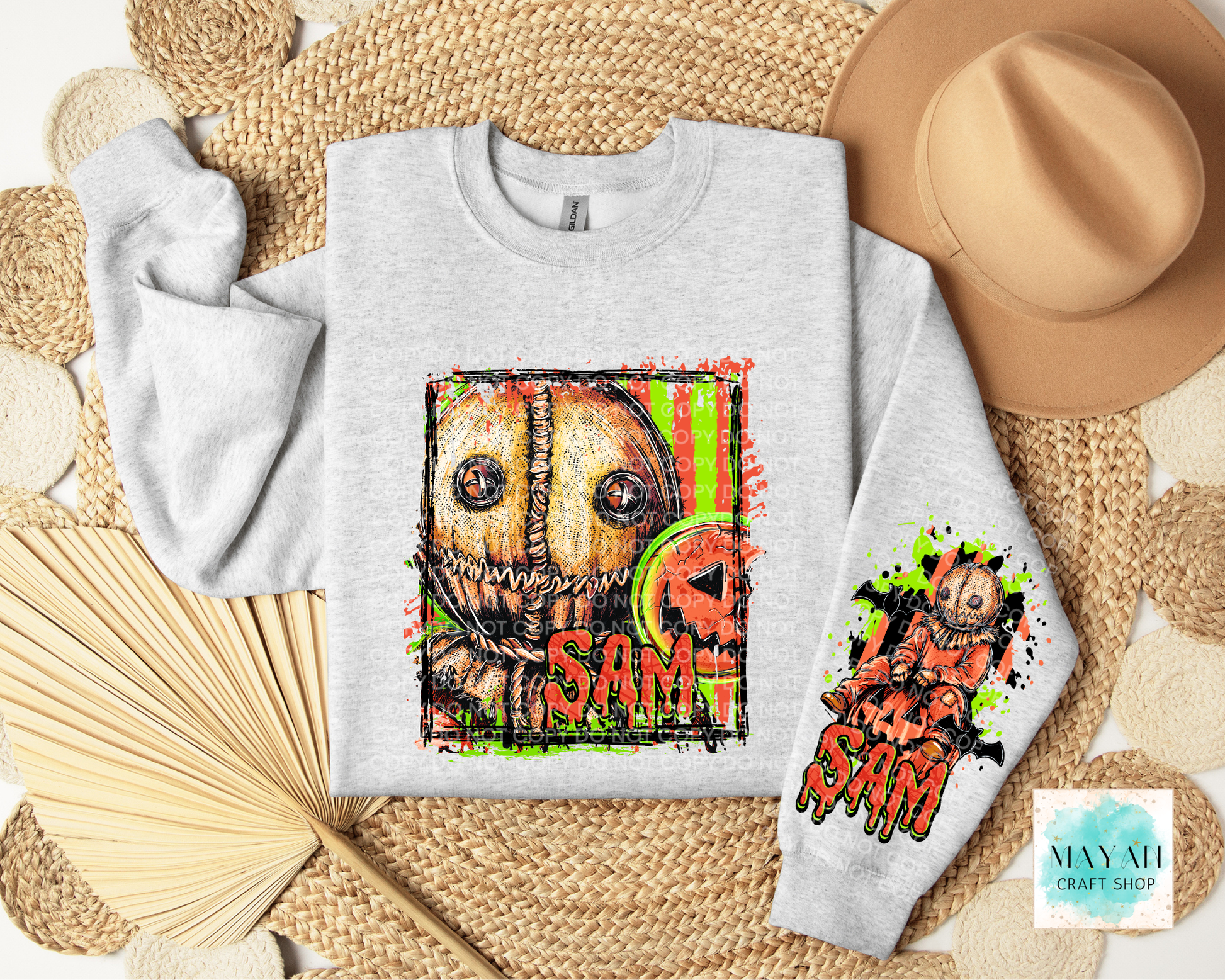 A light gray crewneck sweatshirt featuring a colorful graphic of a creepy straw face in orange dripping text, styled in a flat lay with a tan hat, woven mat.