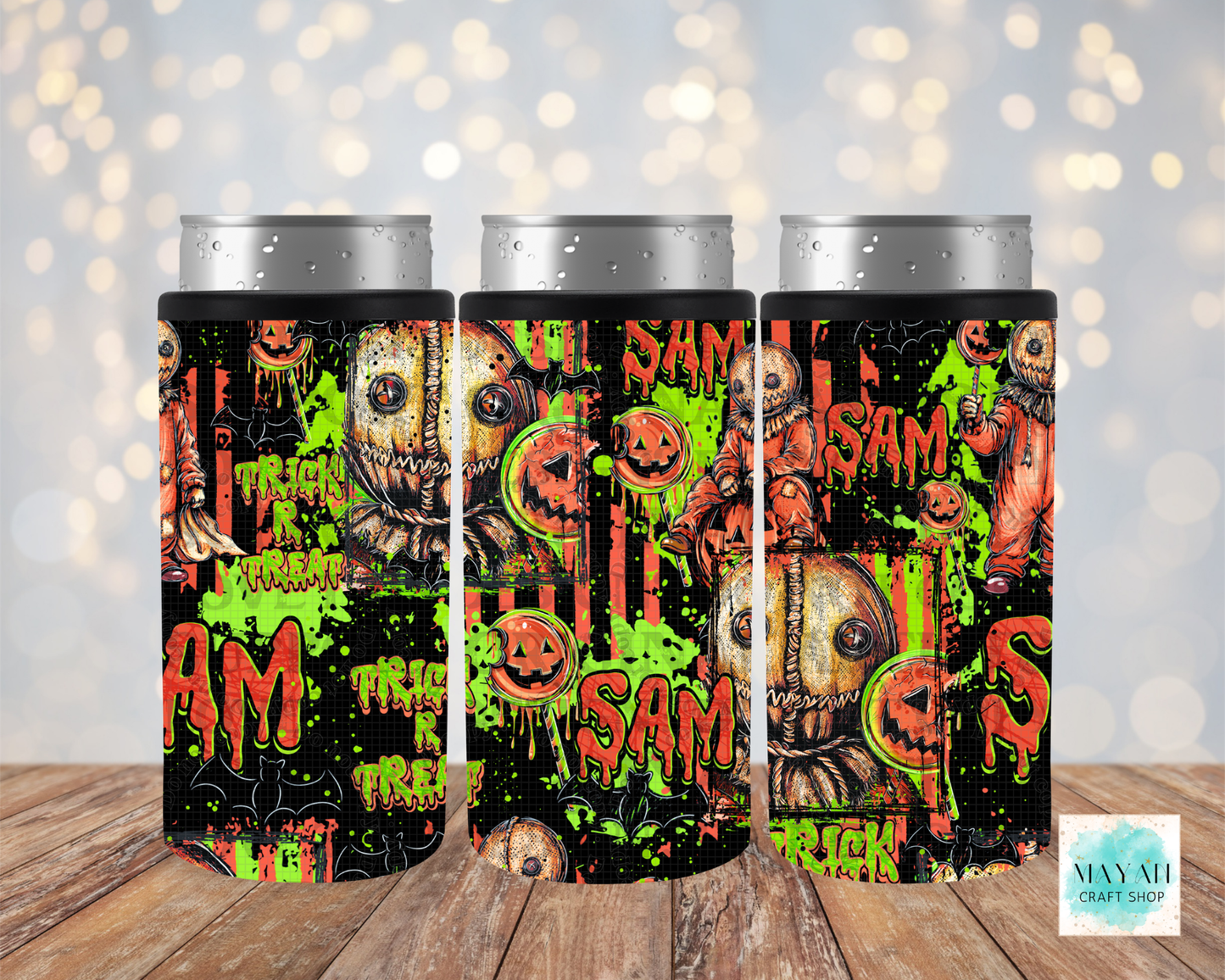 Three slim can coolers with a Halloween-themed collage print featuring pumpkins, skulls, in dripping orange text, shown on silver aluminum cans against a blurred warm bokeh background and wood surface.