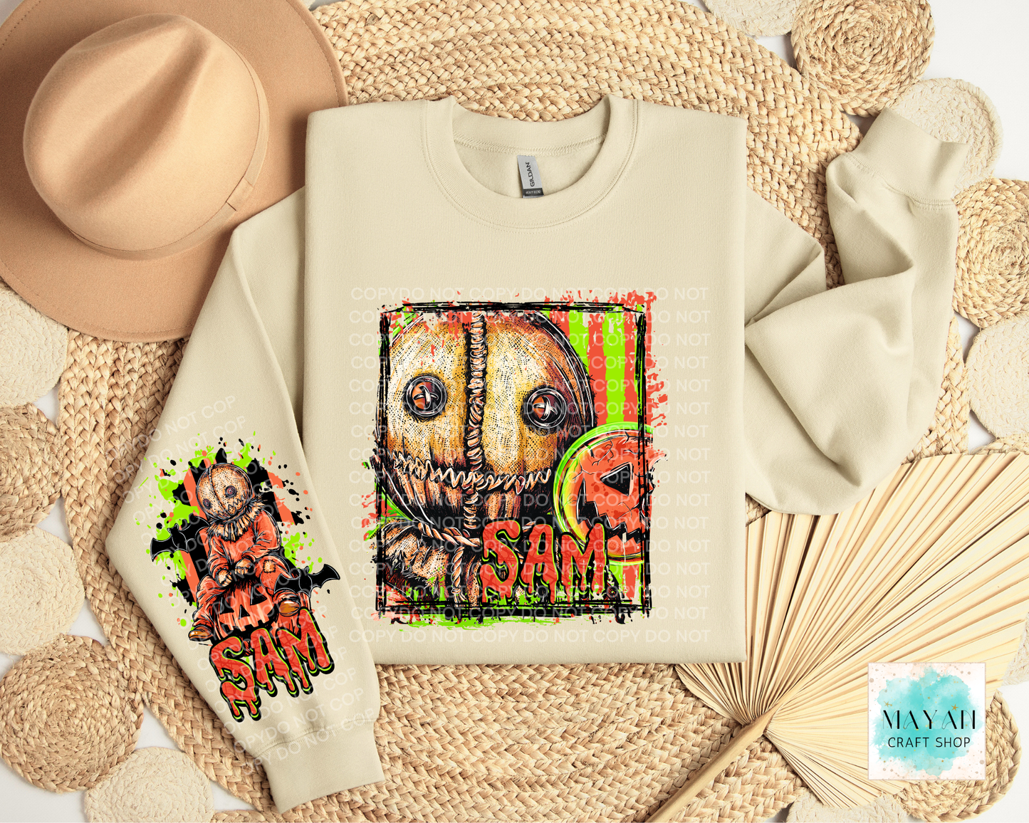 A cream-colored sweatshirt with a horror-themed graphic print, styled in a flat-lay arrangement with a beige hat, woven placemats, and a paper fan. 