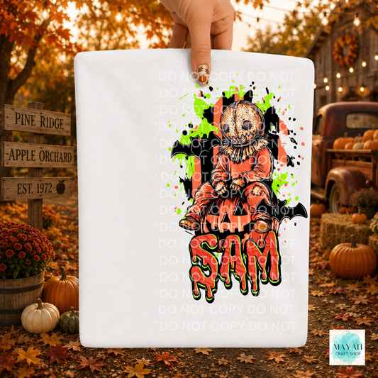 A white rectangular sign or board held by a hand, featuring a Halloween-themed graphic of a scarecrow in orange and black. The scene is set outdoors in a fall yard with pumpkins, autumn leaves, and a rustic truck in the background.