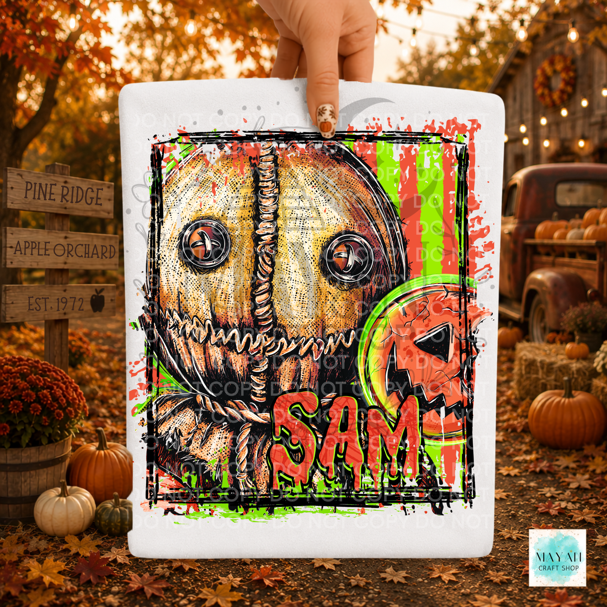 A hand holds a white shirt featuring a Halloween-style illustrated scarecrow face against an autumn outdoor scene with pumpkins, fall leaves, and rustic signs.