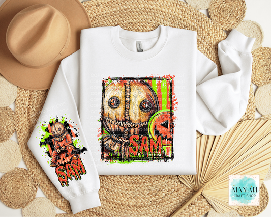 A white crewneck sweatshirt laid flat on a woven straw mat, featuring a large colorful horror-style face graphic on the front and a smaller matching graphic on the left sleeve. A tan hat, woven mat, and dried palm leaves surround it as styling props.