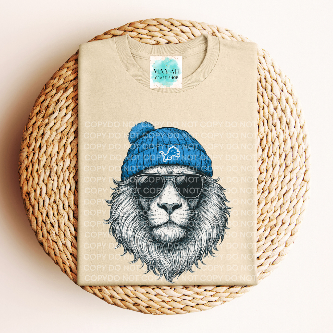 Beige t-shirt with a lion graphic wearing a blue beanie and sunglasses on a woven surface.