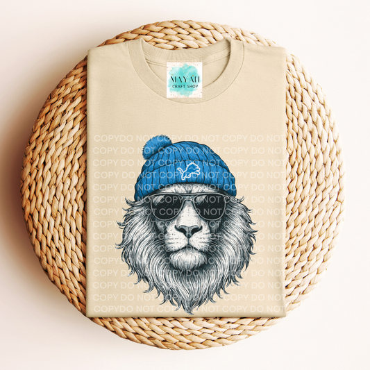 Beige t-shirt with a lion graphic wearing a blue beanie and sunglasses on a woven surface.