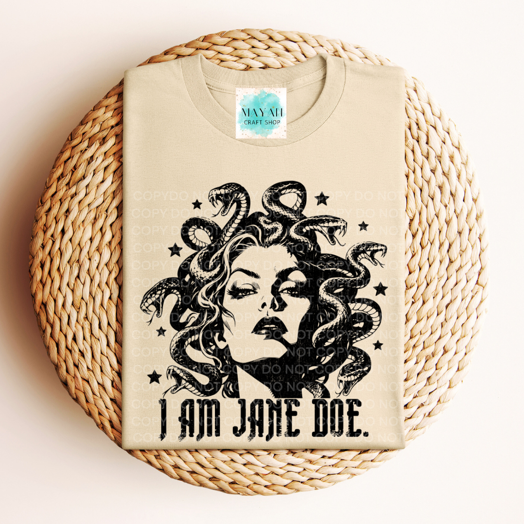 A sand graphic t-shirt featuring a black illustration of Medusa with the text 'I AM JANE DOE.' printed below it, displayed flat on a round woven straw mat.