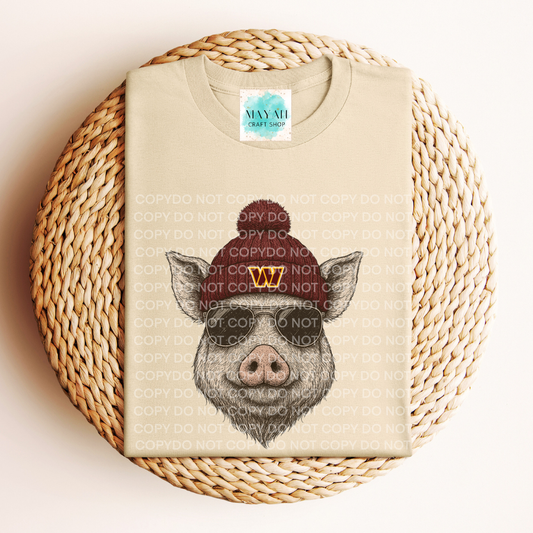 Beige t-shirt with a graphic of a pig wearing a hat on a woven mat