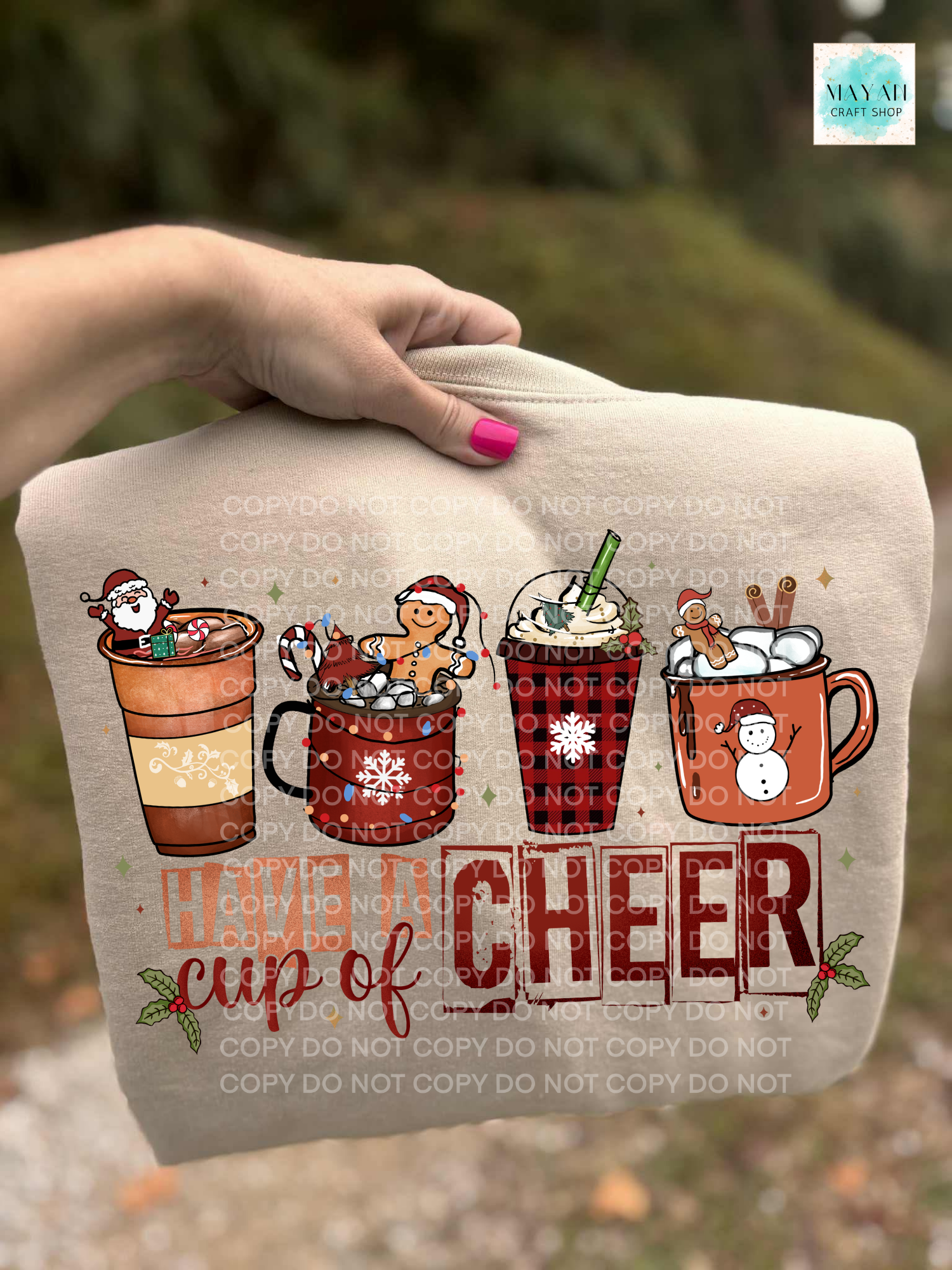Sweatshirt with Christmas-themed design of hot cocoa cups and text 'Have a cup of cheer' held by a hand.
