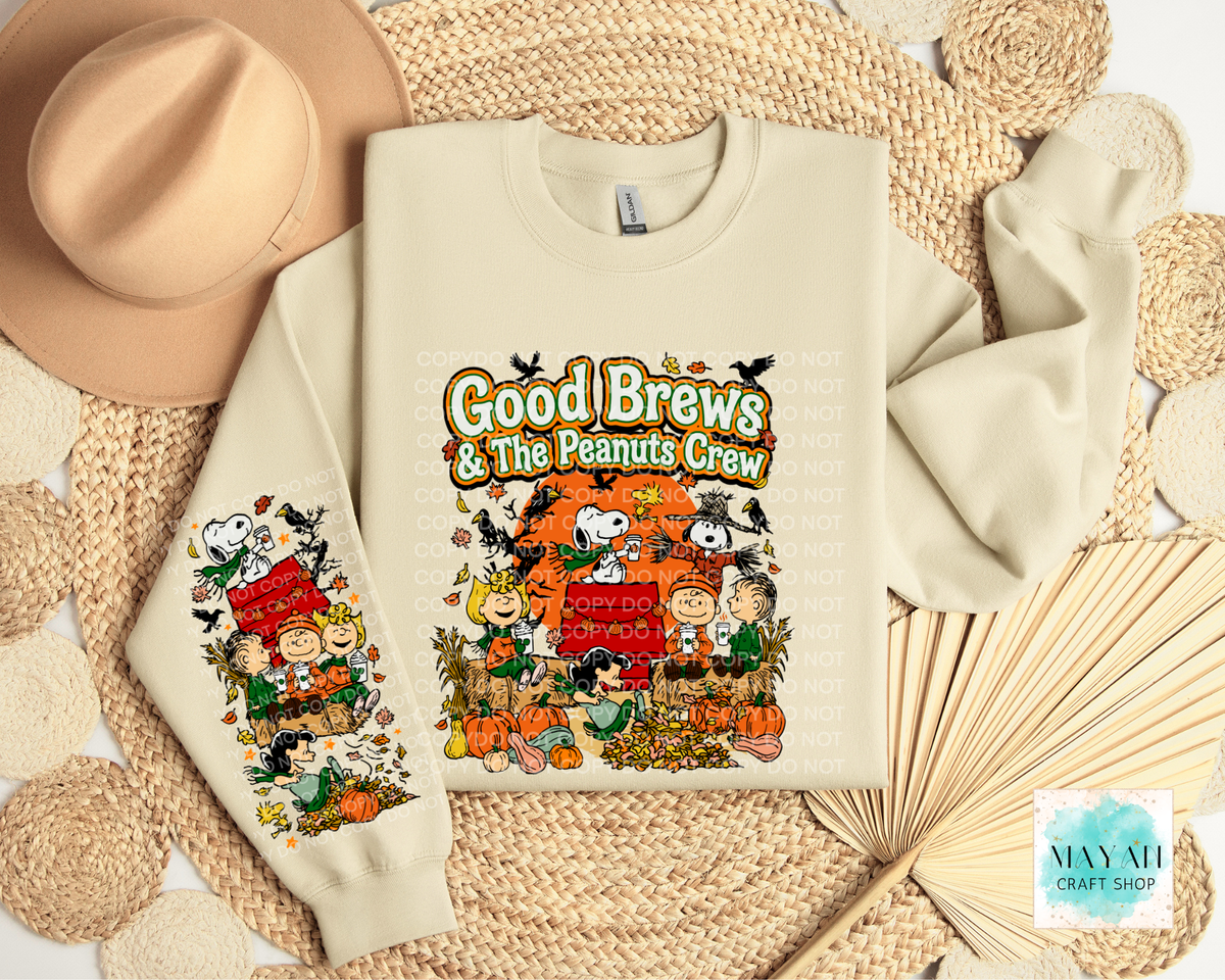 A cream-colored crewneck sweatshirt featuring a colorful 'Good Brews' autumn-themed graphic, styled in a flat lay with a tan hat, woven placemats, and a paper fan.