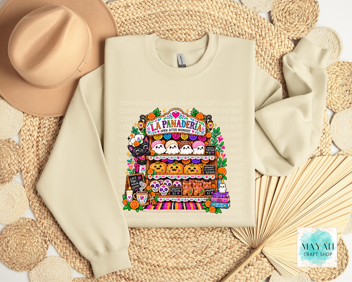 A cream-colored crewneck sweatshirt featuring a colorful graphic of a Halloween-themed stand called 'La Panadería' with cute characters and treats, styled in a flat lay with a beige hat, woven placemat.