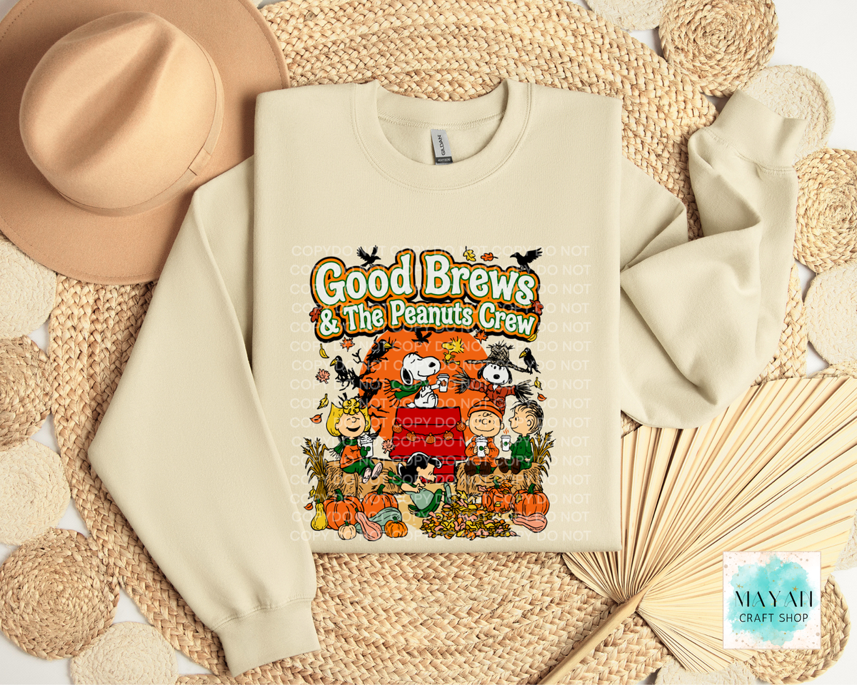 A cream-colored crewneck sweatshirt featuring a colorful 'Good Brews' fall-themed graphic, styled in a flat lay with a tan hat, woven placemat, and paper fan.