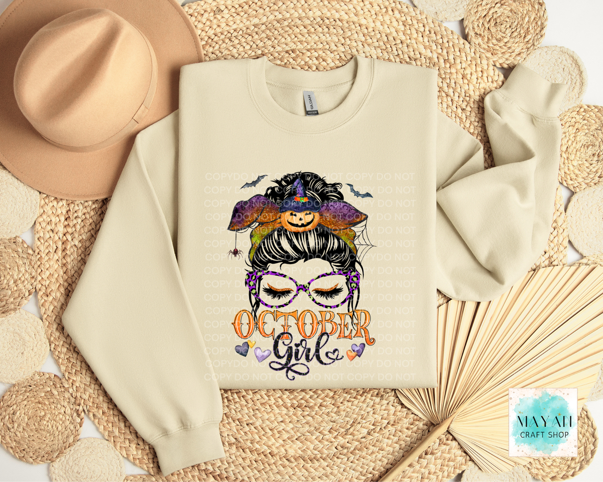 A cream-colored crewneck sweatshirt featuring a colorful graphic of a witch-themed girl with the text 'OCTOBER Girl' underneath, styled in a flat-lay arrangement with a beige hat, woven mat, and paper fan.