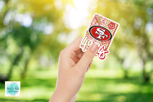 A hand holding a die-cut sticker featuring football team logo in red and white script, set against a blurred green park background with bright sunlight.