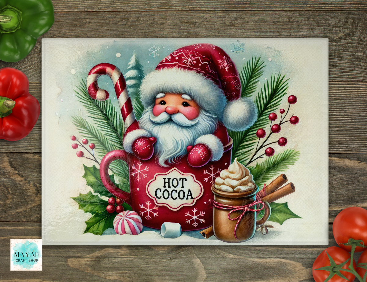 A cutting board featuring a festive Christmas illustration of Santa Claus in a red mug labeled 'HOT COCOA,' surrounded by pine branches, holly, candy canes, and a hot chocolate cup, displayed on a rustic wood surface with decorative tomatoes around the edges.