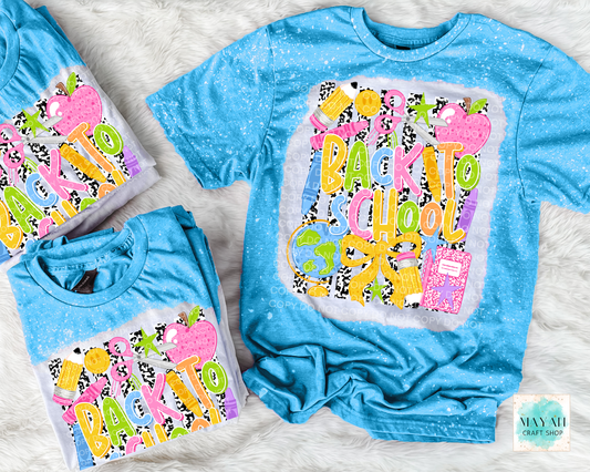 Three blue short-sleeve graphic T-shirts arranged on a white faux-fur surface. The shirts feature a colorful 'Back to School' design with pink apples, yellow and purple lettering, and a pink notebook motif.