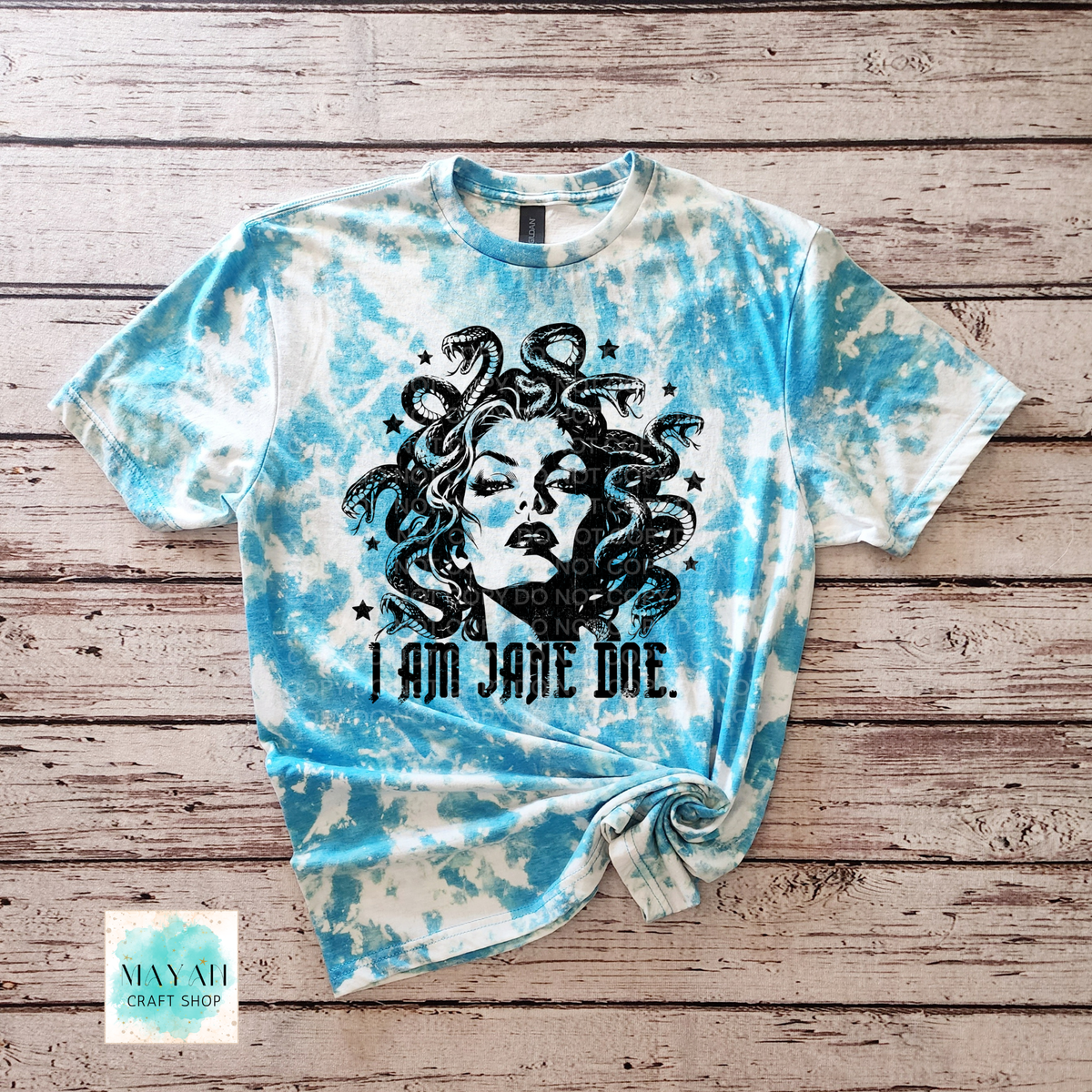 A heather sapphire bleached graphic t-shirt featuring a black illustration of Medusa and the text 'I AM JANE DOE.' laid flat on a rustic wooden plank background.