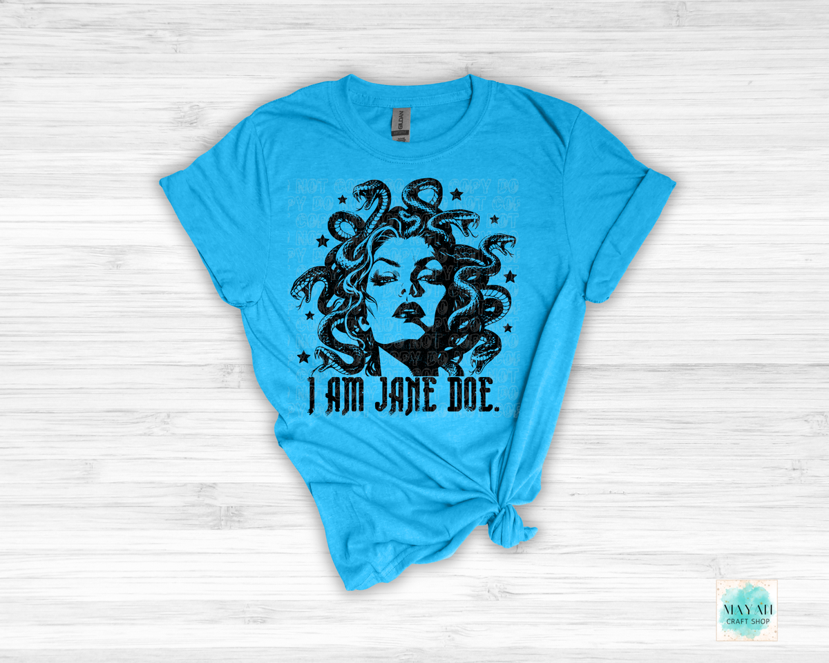 A heather sapphire graphic t-shirt featuring a black print of Medusa with the text 'I AM JANE DOE.' underneath, styled flat on a white wood background with a knot at the bottom hem.