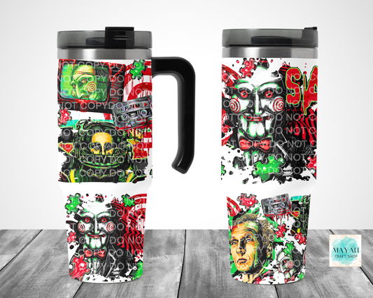 Two insulated travel mugs with black lids and handles, shown from front and side views on a gray wood surface. The mugs feature a bold horror-themed collage print with faces, cassette tapes, and graffiti-style graphics in red, green, black, and white.