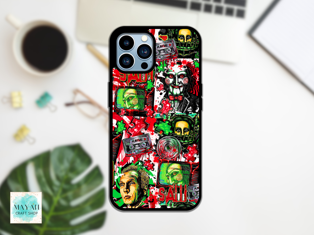 A black phone case with a colorful, chaotic collage design featuring skulls, cassette tapes, faces, and a word in red, styled in a flat-lay on a white desk with a coffee cup, glasses, and a laptop.