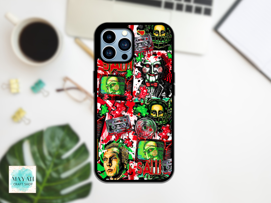 A black phone case with a colorful, chaotic collage design featuring skulls, cassette tapes, faces, and a word in red, styled in a flat-lay on a white desk with a coffee cup, glasses, and a laptop.