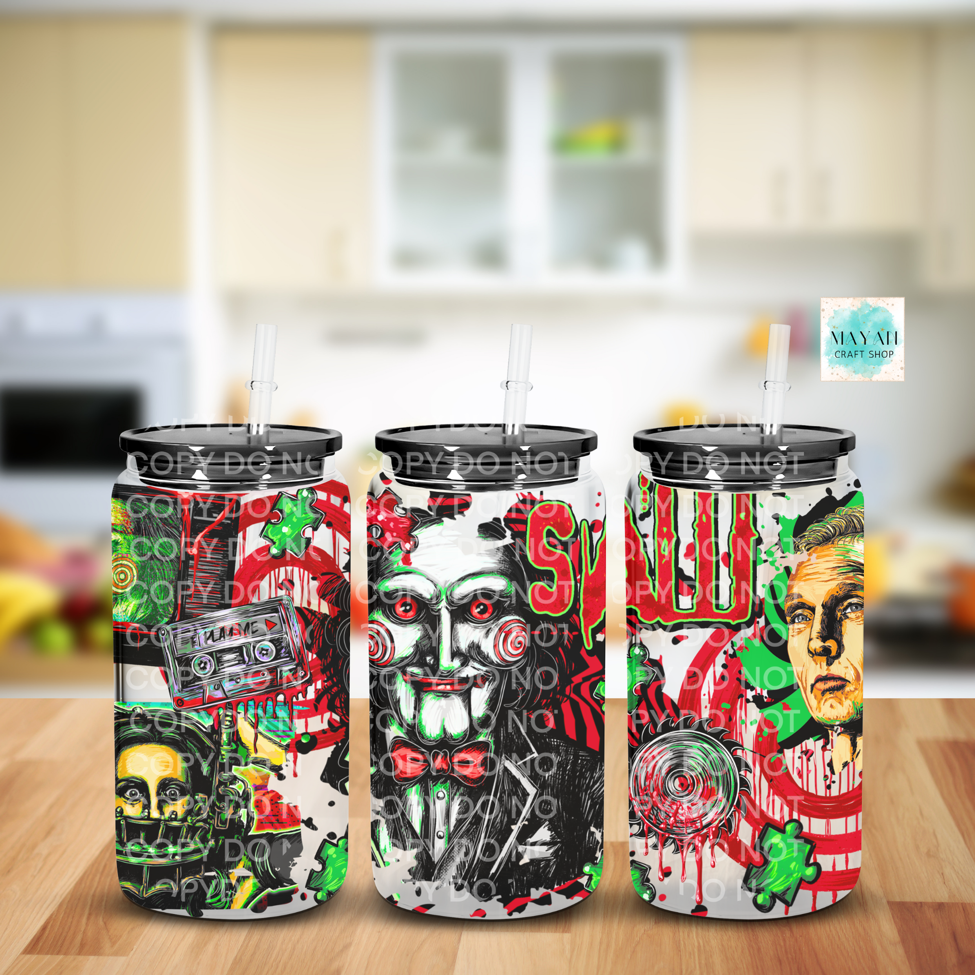 Three tall frosted glass cans with black lids and clear straws, each printed with a different horror-themed graphic design, displayed on a wooden surface with a blurred kitchen background. 