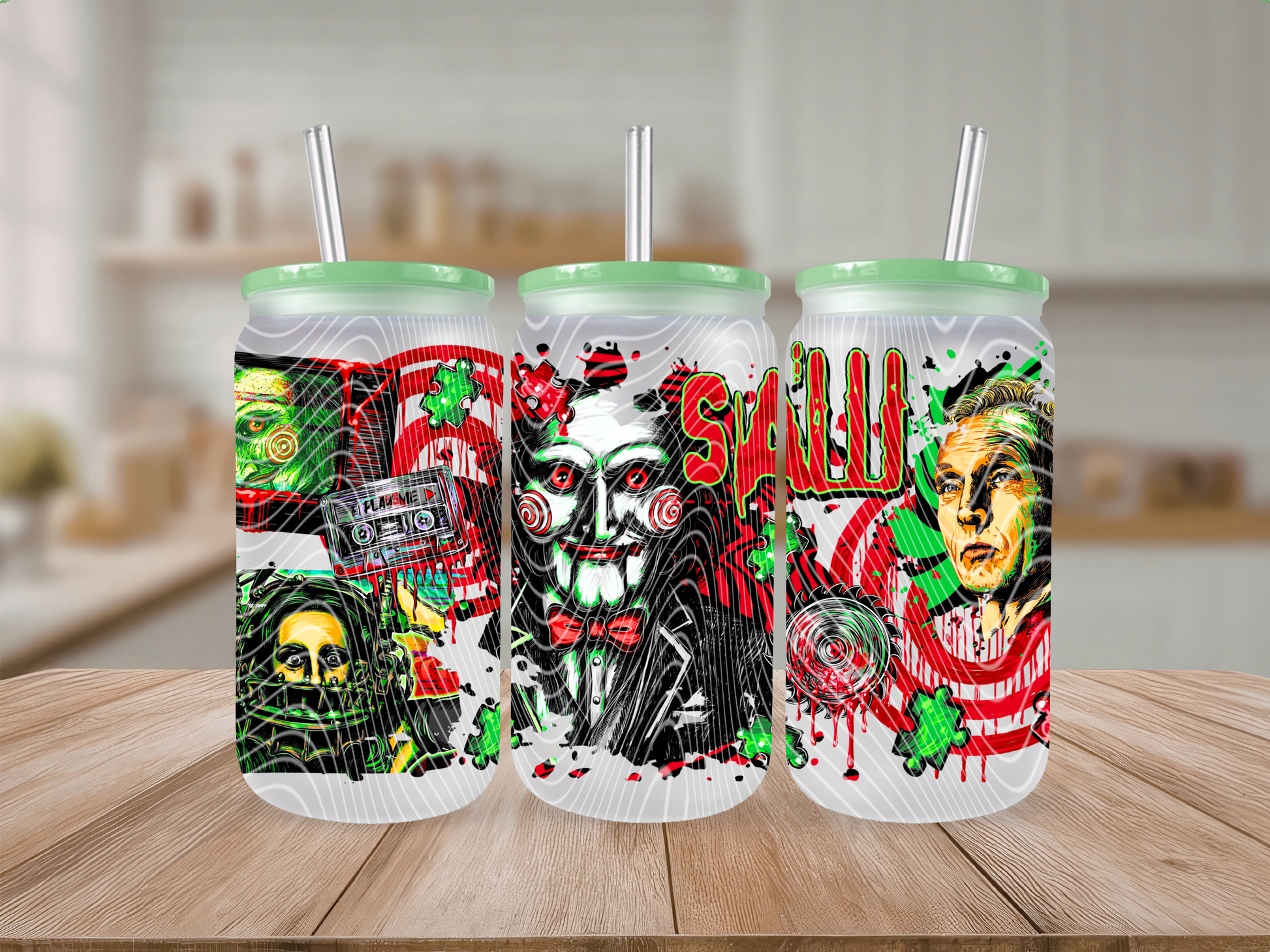 Three tall clear tumblers with mint green lids and white straws, decorated with horror-themed graphics including a skull, a masked face, and a face print, shown on a wooden surface with a blurred indoor background.