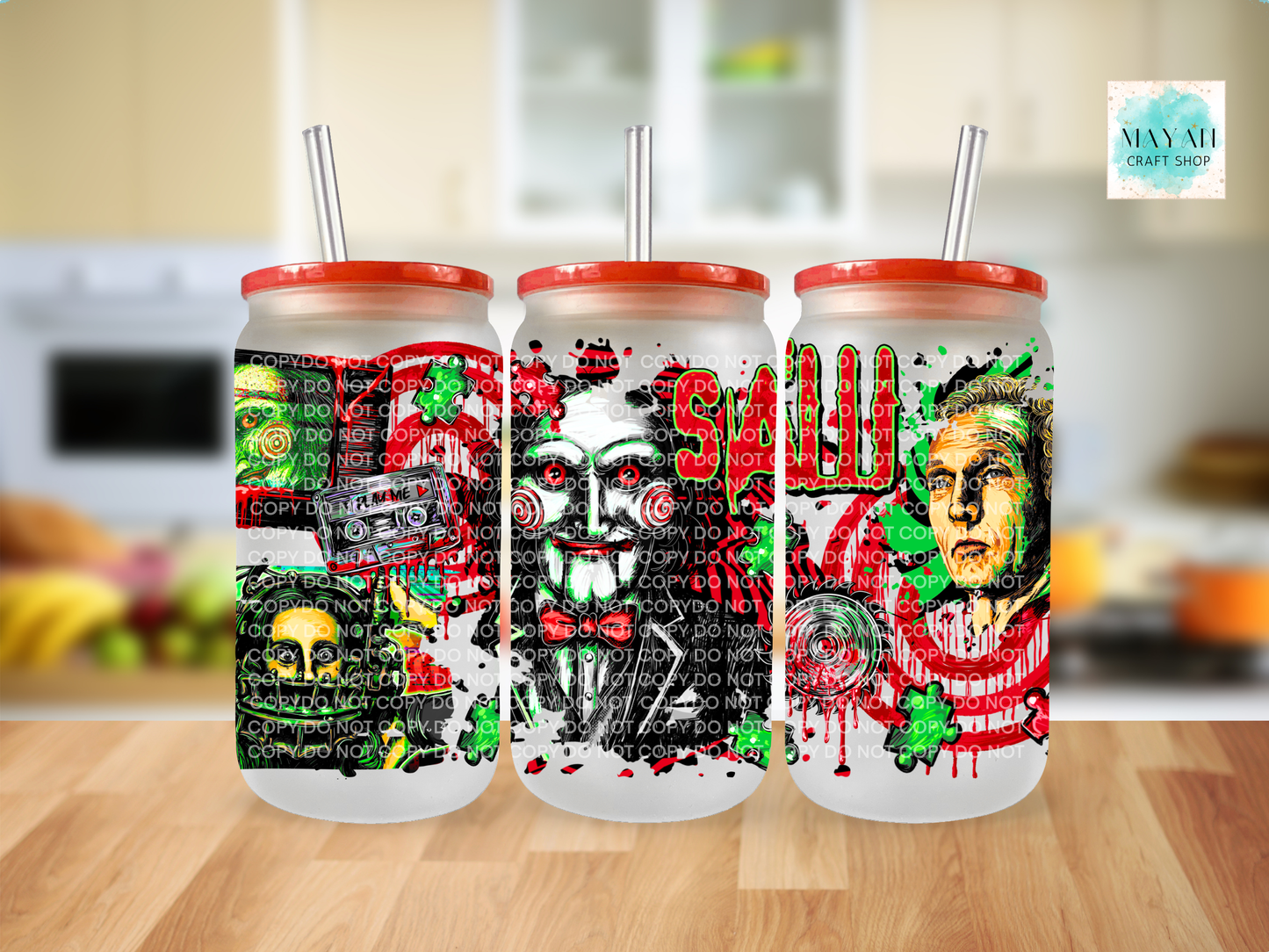 Three frosted glass can-shaped tumblers with red lids and clear straws, featuring horror-themed printed graphics, and a face illustration, displayed on a wooden surface with a blurred kitchen background.