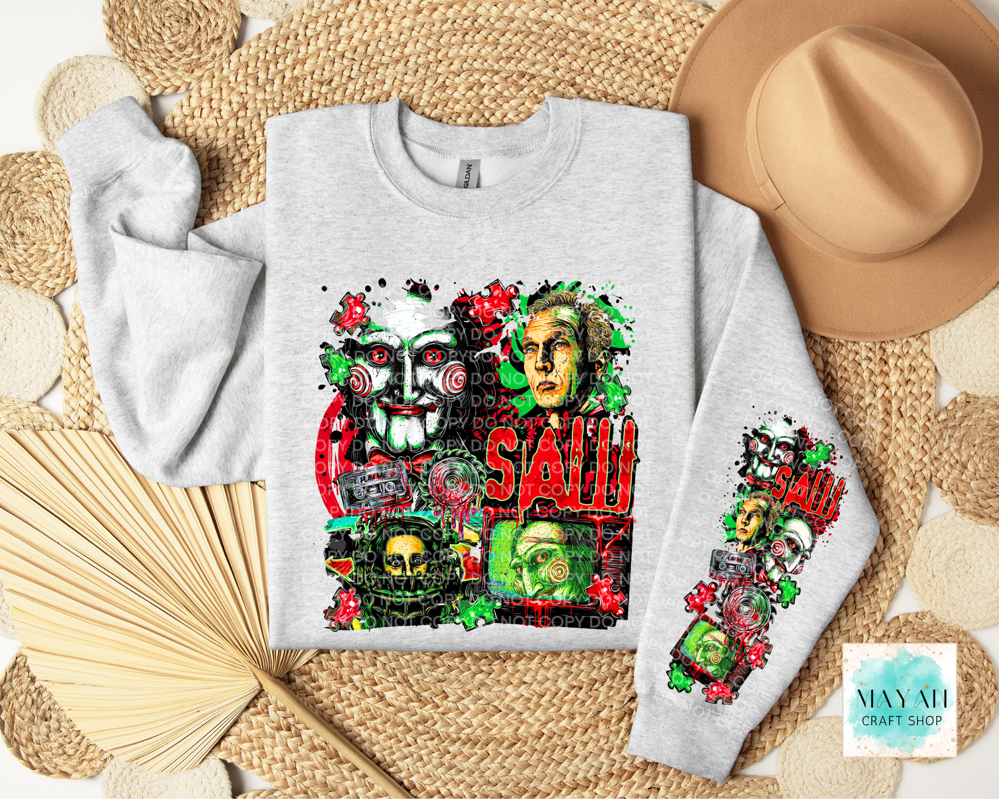 A light gray sweatshirt laid flat on a woven straw mat, decorated with a horror-themed collage print featuring masks, skulls. A tan hat, folded beige towel, and wooden fan are arranged around it.